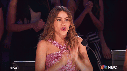 A Woman In A Pink Dress Is Clapping In Front Of A Microphone With The Nbc Logo In The Background GIF