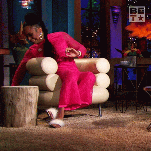 A Woman In A Pink Dress Is Sitting In A Chair With A Sign That Says Be GIF