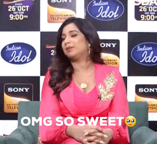 A Woman In A Pink Dress Is Sitting In Front Of A Wall With Indian Idol Logos On It GIF