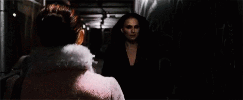 A Woman In A Pink Dress Is Standing In A Dark Room Looking At Another Woman . GIF