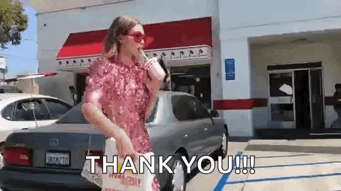 A Woman In A Pink Dress Is Standing In Front Of A Car Holding A Cup Of Coffee And A Bag Of Food . GIF