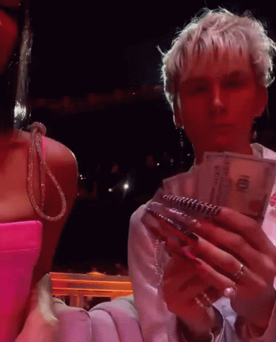 A Woman In A Pink Dress Is Standing Next To A Man In A White Shirt Holding A Stack Of 100 Dollar Bills GIF