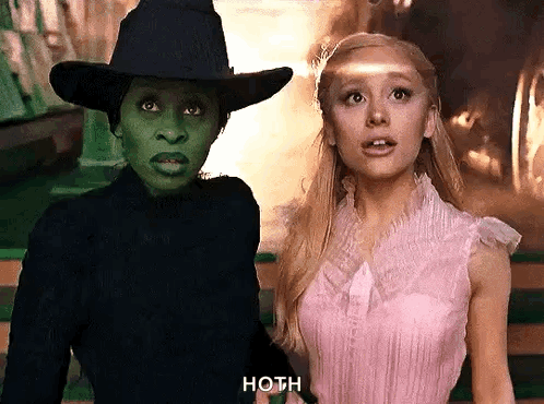 A Woman In A Pink Dress Is Standing Next To A Witch In A Black Hat And Mask GIF