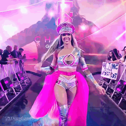 A Woman In A Pink Dress Is Walking Down A Runway With A Sign That Says Waiting Is The Hardest Part GIF
