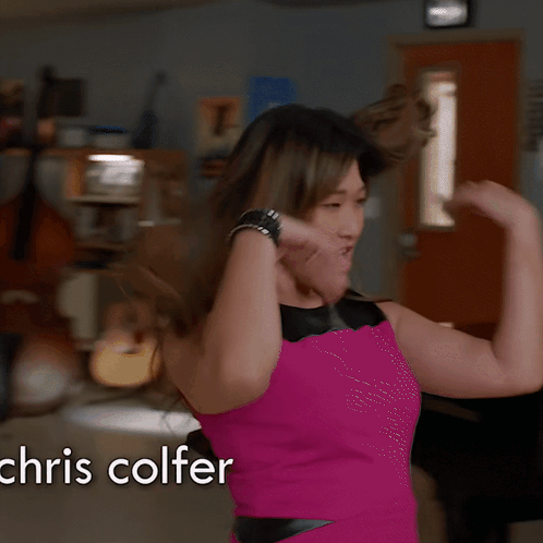 A Woman In A Pink Dress Stands In Front Of A Piano With The Name Chris Colfer On The Bottom GIF
