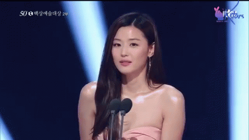 A Woman In A Pink Dress Stands In Front Of Two Microphones With A Heart On The Bottom Right Corner GIF