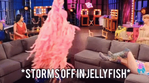 A Woman In A Pink Feathered Dress Is Standing In Front Of A Couch And Says Storms Off In Jellyfish . GIF
