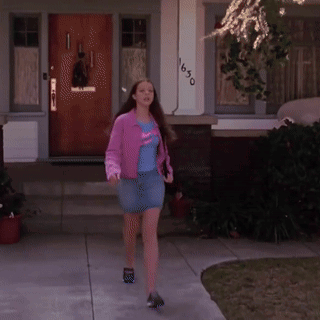 A Woman In A Pink Jacket And Blue Skirt Is Walking In Front Of A House With The Number 1650 On It GIF