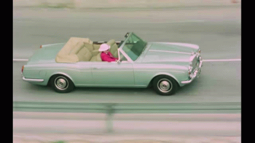 A Woman In A Pink Jacket Is Driving A Silver Convertible GIF