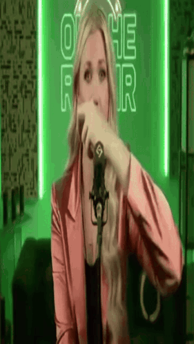 A Woman In A Pink Jacket Is Singing Into A Microphone In Front Of A Green Background . GIF