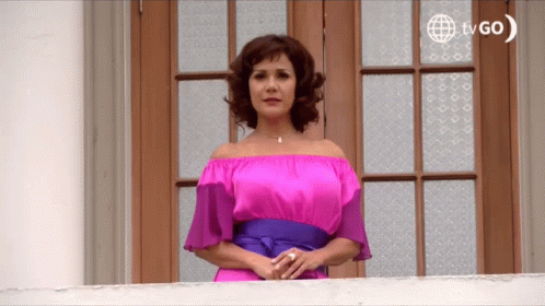 A Woman In A Pink Off The Shoulder Dress Stands On A Balcony With A Tvgo Logo In The Background GIF