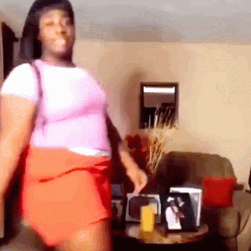 A Woman In A Pink Shirt And Orange Skirt Is Standing In A Living Room . GIF