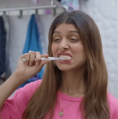 A Woman In A Pink Shirt Is Brushing Her Teeth With A Toothbrush GIF