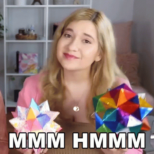 A Woman In A Pink Shirt Is Holding A Colorful Origami Object And Says Hmm GIF