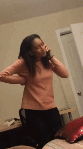 A Woman In A Pink Shirt Is Standing In A Room Talking On A Phone GIF
