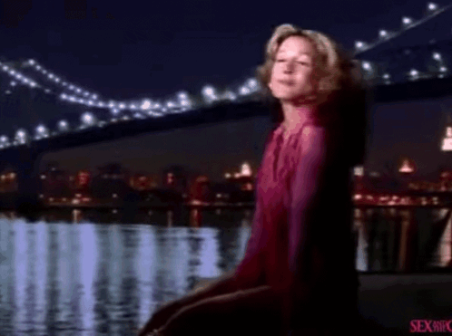 A Woman In A Pink Shirt Is Standing In Front Of A Body Of Water And A Bridge . GIF