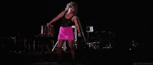 A Woman In A Pink Skirt Is Singing Into A Microphone On Stage . GIF