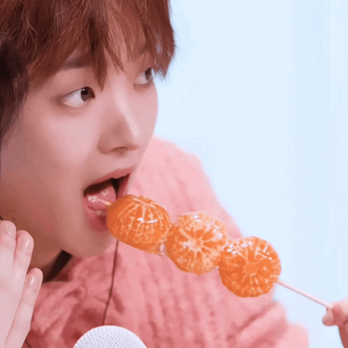 A Woman In A Pink Sweater Is Eating A Skewer Of Oranges On A Stick GIF