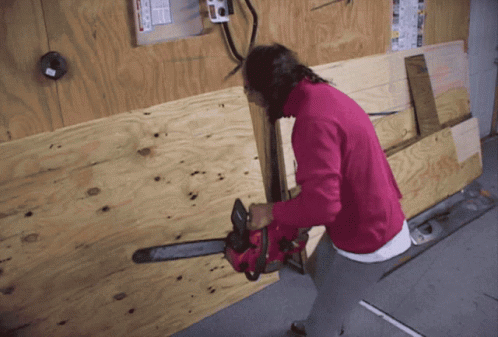 A Woman In A Pink Sweater Is Holding A Chainsaw Against A Wooden Wall GIF
