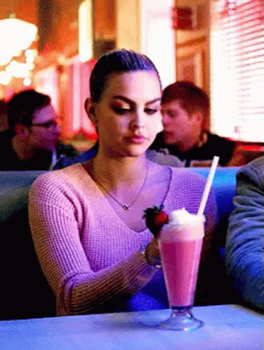 A Woman In A Pink Sweater Is Sitting At A Table With A Milkshake And A Strawberry GIF