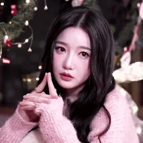 A Woman In A Pink Sweater Is Sitting In Front Of A Christmas Tree With Her Hands Folded . GIF