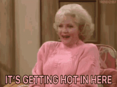 A Woman In A Pink Sweater Is Sitting On A Couch And Says It 'S Getting Hot In Here . GIF