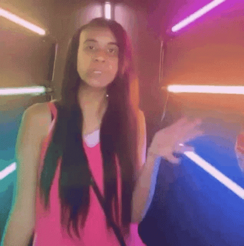 A Woman In A Pink Tank Top Is Standing In Front Of A Neon Sign . GIF