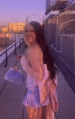 A Woman In A Pink Top And Blue Skirt Is Standing On A Balcony Holding A Purple Purse . GIF