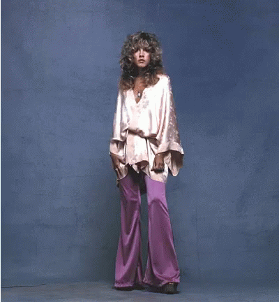A Woman In A Pink Top And Purple Pants Stands In Front Of A Blue Background GIF