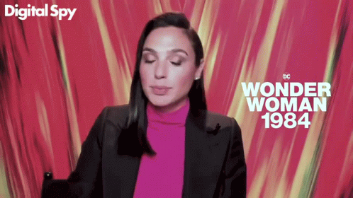 A Woman In A Pink Turtleneck And Black Jacket Is Sitting In Front Of A Digital Spy Wonder Woman 1984 Poster GIF