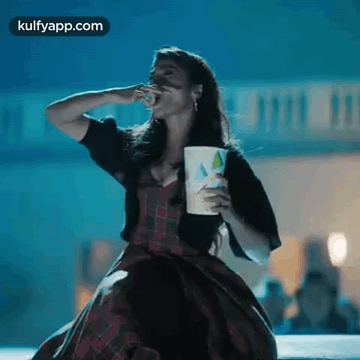 A Woman In A Plaid Dress Is Eating Popcorn And Holding A Cup . GIF