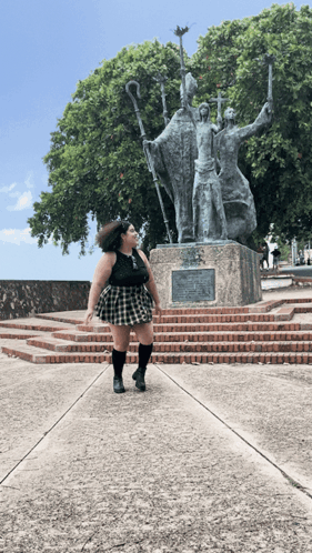 A Woman In A Plaid Skirt Stands In Front Of A Statue Of Three Men GIF