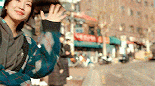 A Woman In A Plaid Sweater Is Waving Her Hand In Front Of A Building . GIF