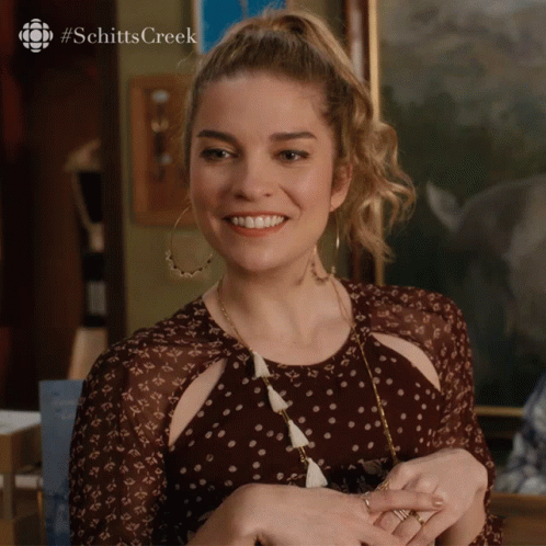 A Woman In A Polka Dot Dress Is Smiling With The #Schittscreek Logo Behind Her GIF
