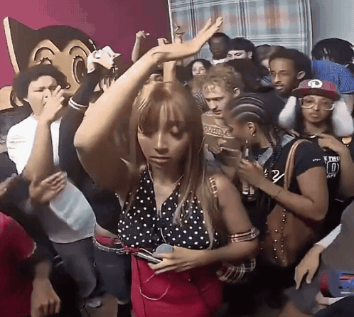 A Woman In A Polka Dot Top Is Dancing In A Crowd With A Man Wearing A Hat With The Letter O On It GIF