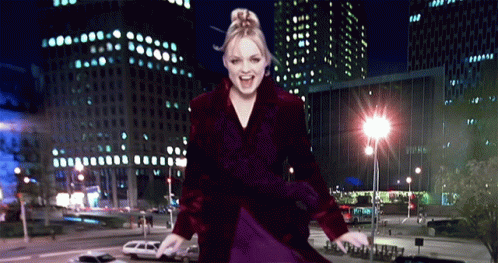 A Woman In A Purple Coat Is Dancing In Front Of A City At Night GIF