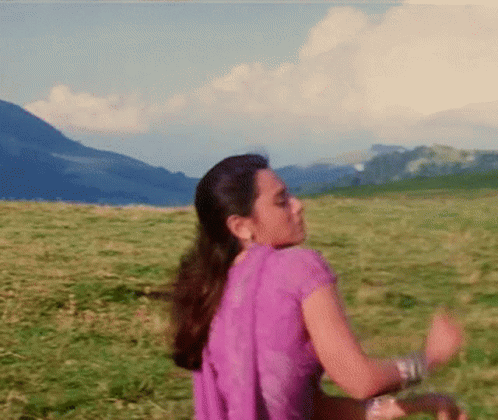 A Woman In A Purple Dress Is Dancing In A Grassy Field With Mountains In The Background . GIF