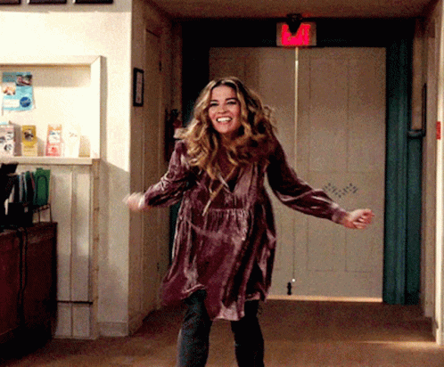A Woman In A Purple Dress Is Dancing In A Hallway With A Red Exit Sign Above Her GIF