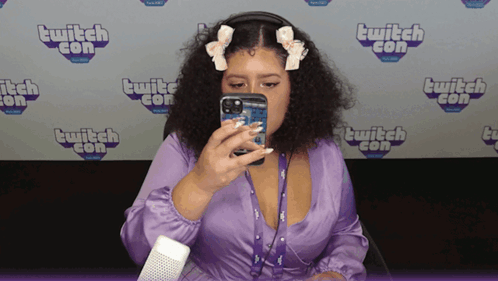 A Woman In A Purple Dress Is Taking A Picture Of Herself In Front Of A Wall That Says Twitch Con GIF