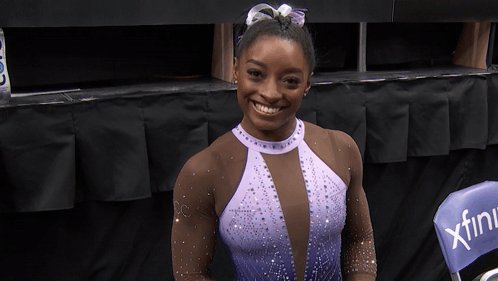 A Woman In A Purple Leotard Is Smiling In Front Of A Chair That Says Xfinity GIF