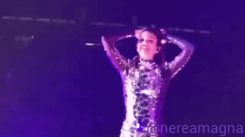 A Woman In A Purple Sequined Dress Is Standing On A Stage In Front Of A Microphone .. GIF