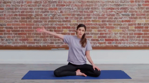 A Woman In A Purple Shirt Sits On A Blue Yoga Mat In Front Of A Brick Wall GIF