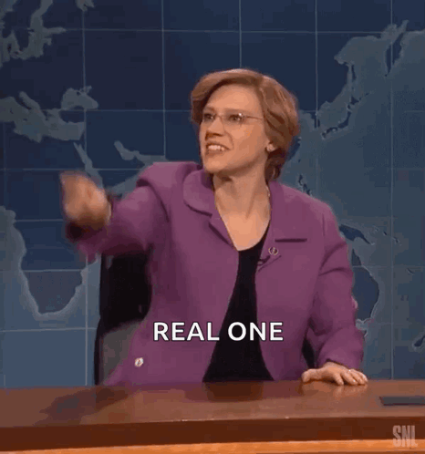 A Woman In A Purple Suit Is Sitting At A Desk And Pointing At The Camera . GIF