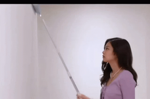 A Woman In A Purple Sweater Is Standing Next To A White Wall And Holding A Vacuum Cleaner . GIF