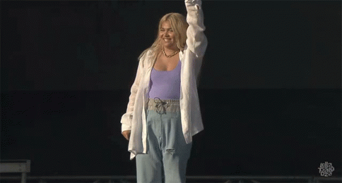 A Woman In A Purple Tank Top And White Shirt Stands On A Stage GIF