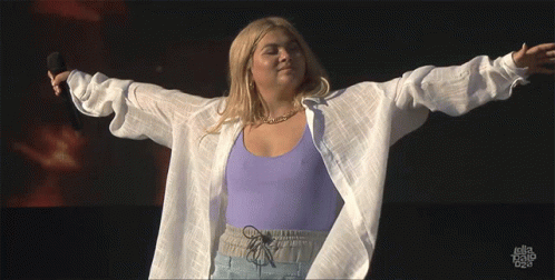 A Woman In A Purple Top And White Shirt Is Holding A Microphone With Her Arms Outstretched GIF