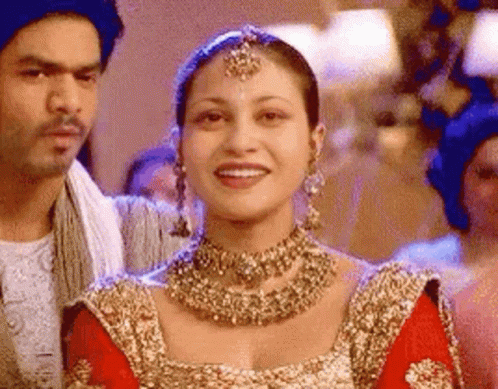 A Woman In A Red And Gold Dress Is Smiling While Standing Next To A Man . GIF