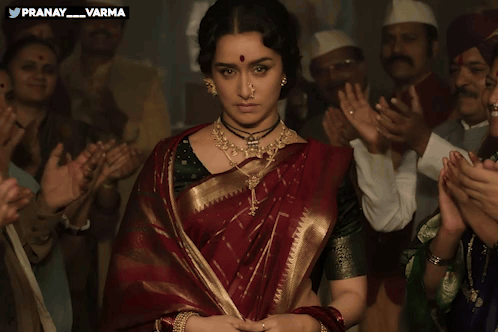 A Woman In A Red And Gold Saree Stands In Front Of A Crowd Of People With The Words Pranay Varma Above Her GIF