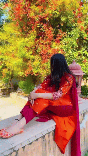 A Woman In A Red And Orange Dress Is Sitting On A Wall . GIF