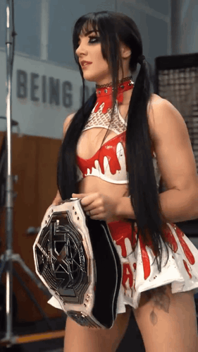 A Woman In A Red And White Outfit Is Holding A Wrestling Belt In Front Of A Sign That Says Being GIF